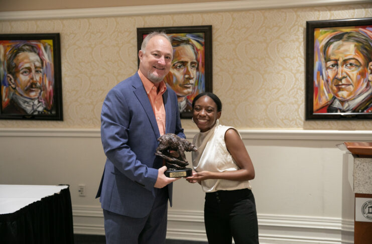 Two people stand indoors holding a trophy together, with framed portraits visible on the wall behind them.