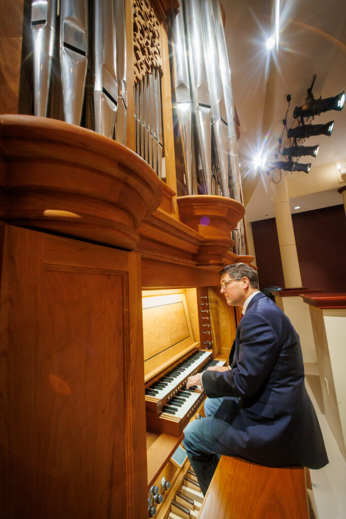 A person in a suit plays a pipe organ in a concert hall with large silver pipes and wooden paneling.