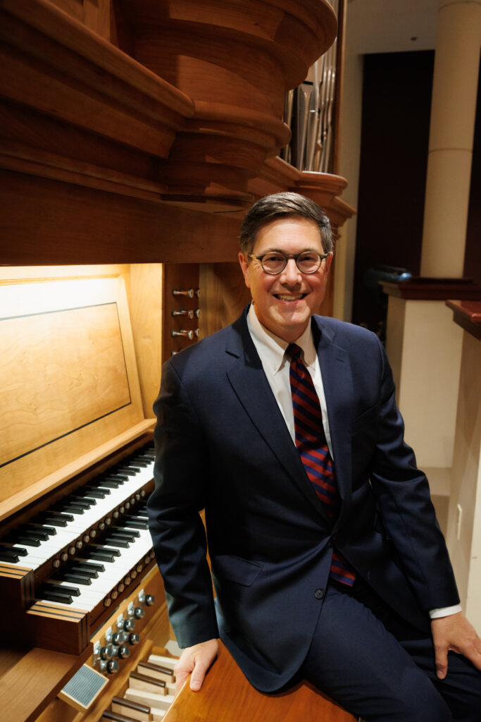 A man in a suit sits beside a large wooden pipe organ, smiling at the camera in a well-lit room.