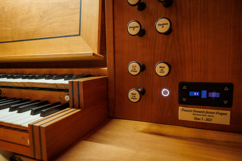 Close-up of a pipe organ console showing keys, stop knobs, a digital display, and a maker’s label.