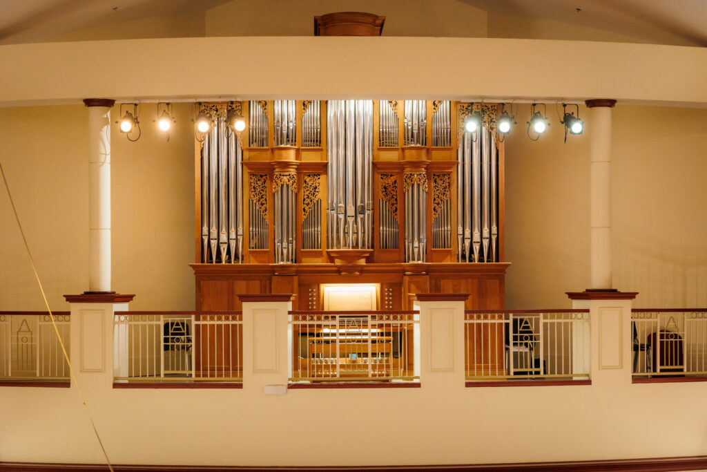 A large pipe organ with ornate woodwork is displayed behind a railing in a well-lit concert hall.