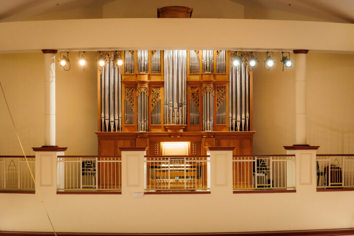 250825MUFicklingFacultyConcert_0027 A large pipe organ with ornate woodwork is displayed behind a railing in a well-lit concert hall.