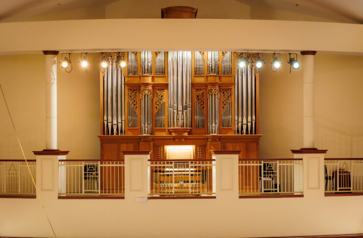 A large pipe organ with ornate woodwork is displayed behind a railing in a well-lit concert hall.