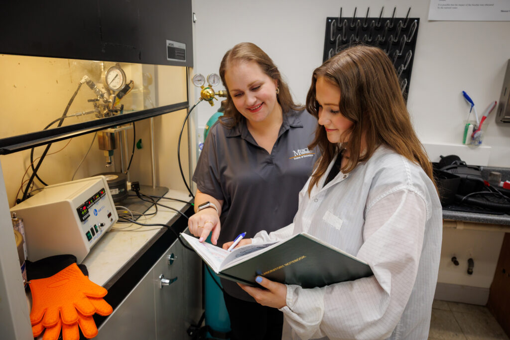 Two women in a laboratory setting review notes in a binder next to lab equipment and safety gloves.