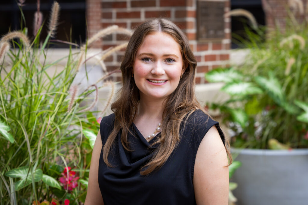 Young woman with long brown hair wearing a black top, smiling outdoors in front of greenery and a brick wall.