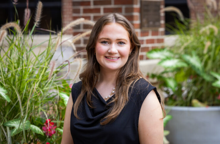 Young woman with long brown hair wearing a black top, smiling outdoors in front of greenery and a brick wall.