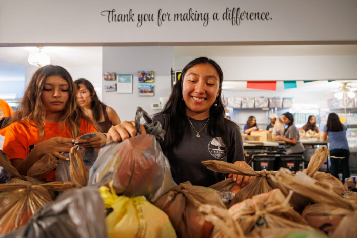 250905MUMerServBackpack_0023 Mercer students pack grocery bags with food in a community center, with a sign reading “Thank you for making a difference.”.