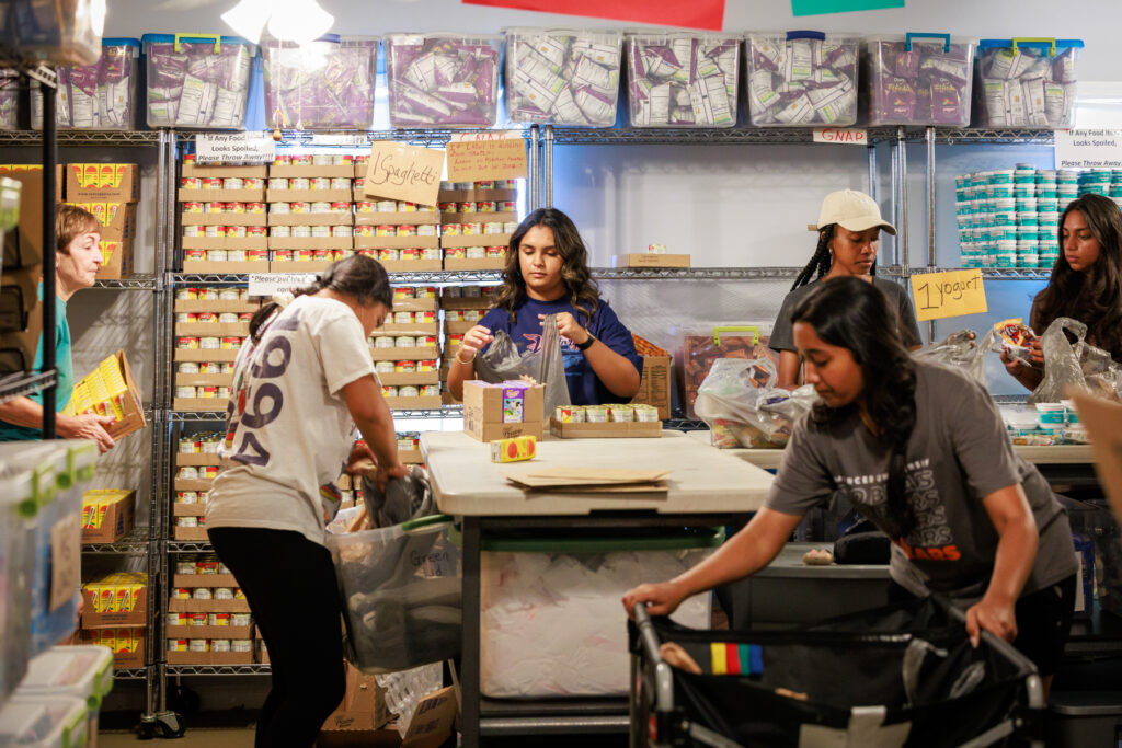 Several people sort and pack food items in a storage room lined with shelves filled with canned goods and supplies.