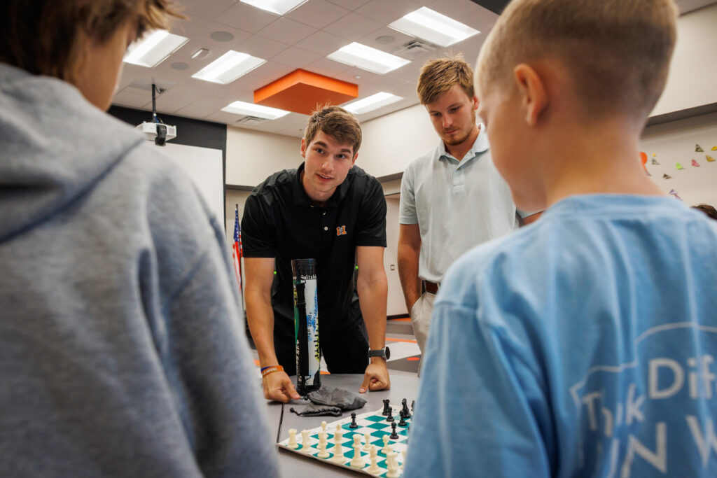 Two Mercer students and two children stand around a chessboard in a classroom, focused on the game.
