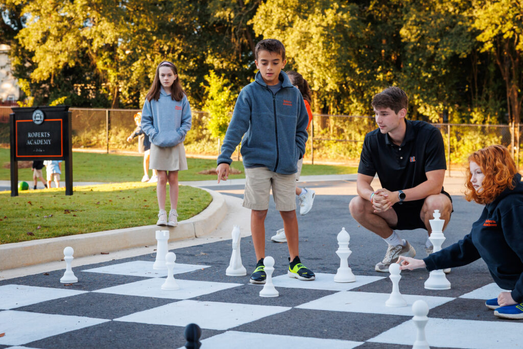 Children and a Mercer student play chess on a large outdoor board, with trees and a school sign visible in the background.