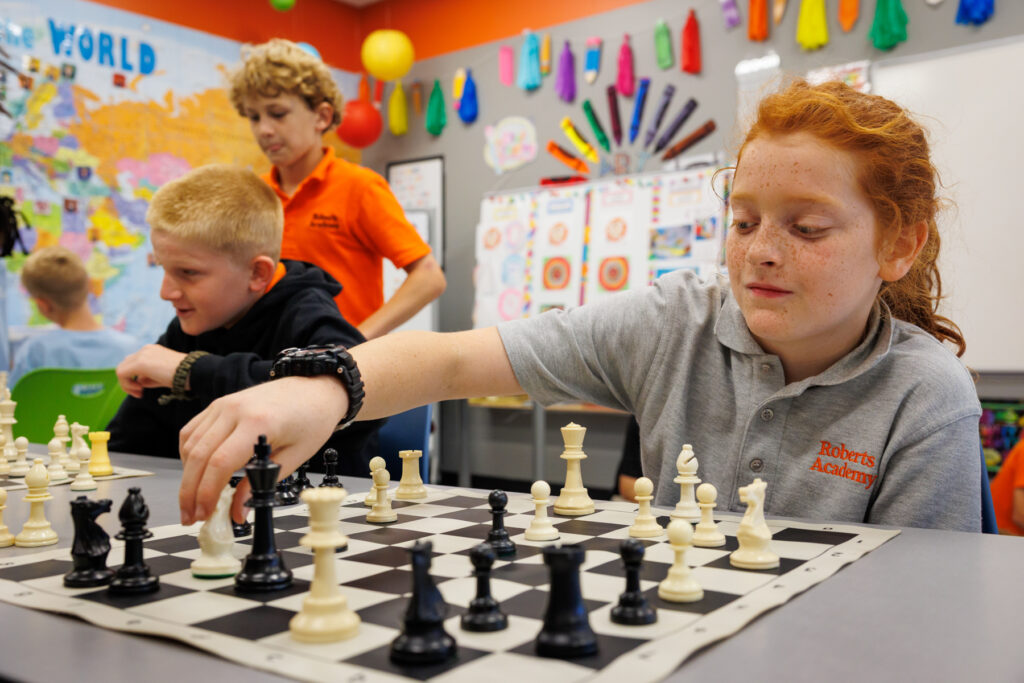 Children play chess in a colorful classroom; one girl in front moves a piece while others play in the background.