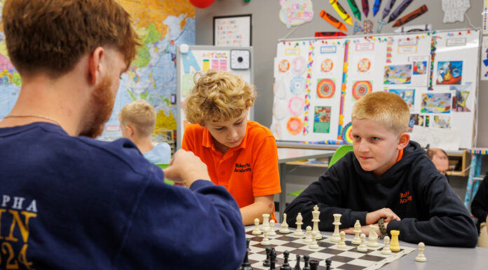 Mercer chess team mentors students at Roberts Academy Two boys and a Mercer student sit at a desk playing chess in a classroom decorated with colorful posters and a world map.
