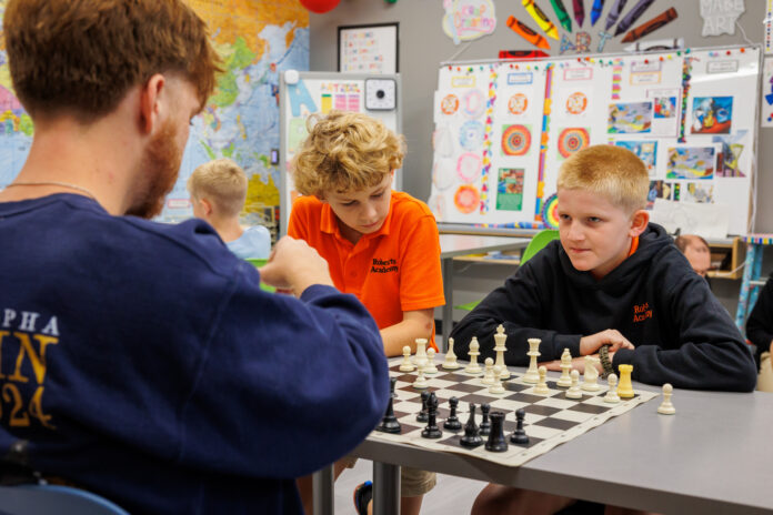 250905MURobertsAcademyChessLY-4 Two boys and a Mercer student sit at a desk playing chess in a classroom decorated with colorful posters and a world map.