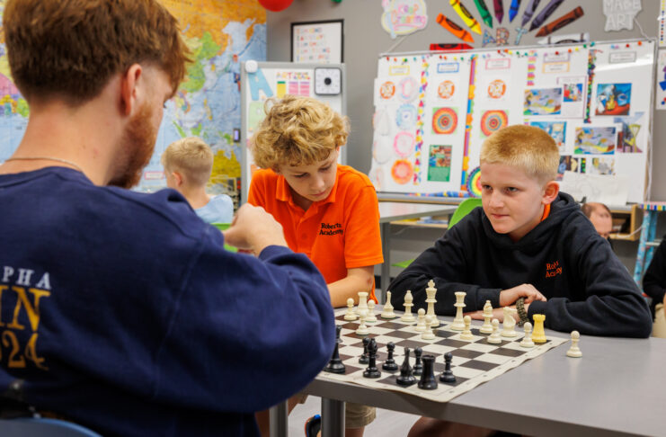 Two boys and a Mercer student sit at a desk playing chess in a classroom decorated with colorful posters and a world map.
