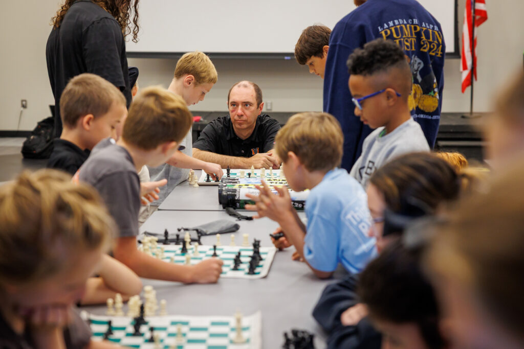 A group of children and an adult sit at tables playing chess in a classroom setting.