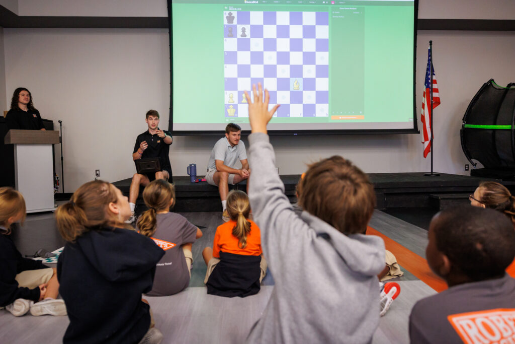 Children sit on the floor facing a screen with a chess game; one child raises a hand to ask a question.