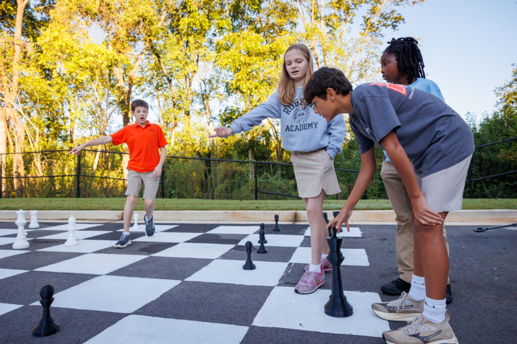 Four children play chess on a large outdoor chessboard, moving oversized chess pieces under bright daylight.
