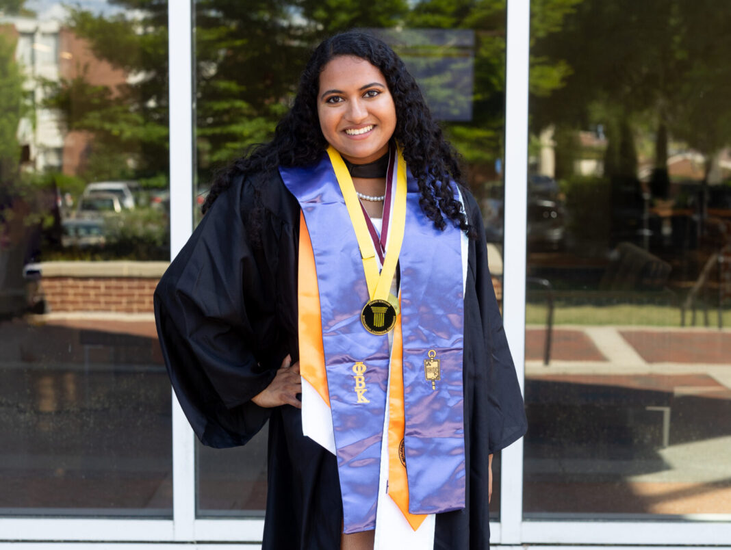 Arsha Moorthy-Tweedie A graduate in cap and gown with honor cords and medals stands outside in front of a glass building, smiling at the camera.