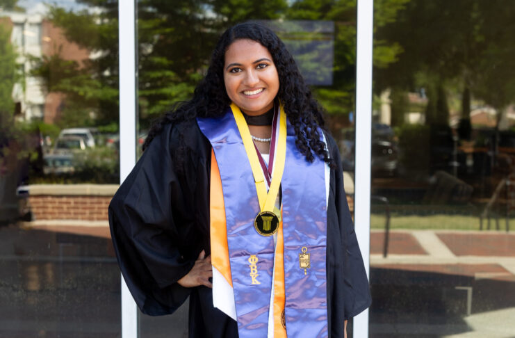 A graduate in cap and gown with honor cords and medals stands outside in front of a glass building, smiling at the camera.