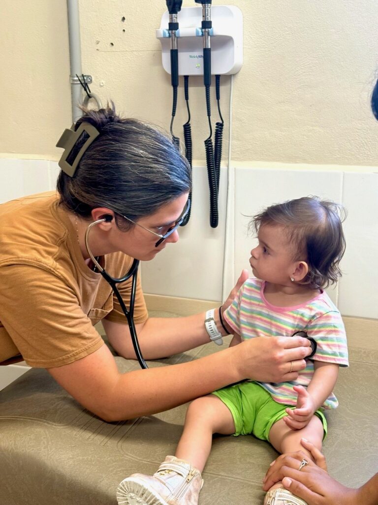 A doctor uses a stethoscope to examine a toddler sitting on an exam table in a medical office.