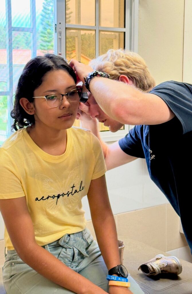 A person examines a girls ear with an otoscope in a brightly lit room near a window.
