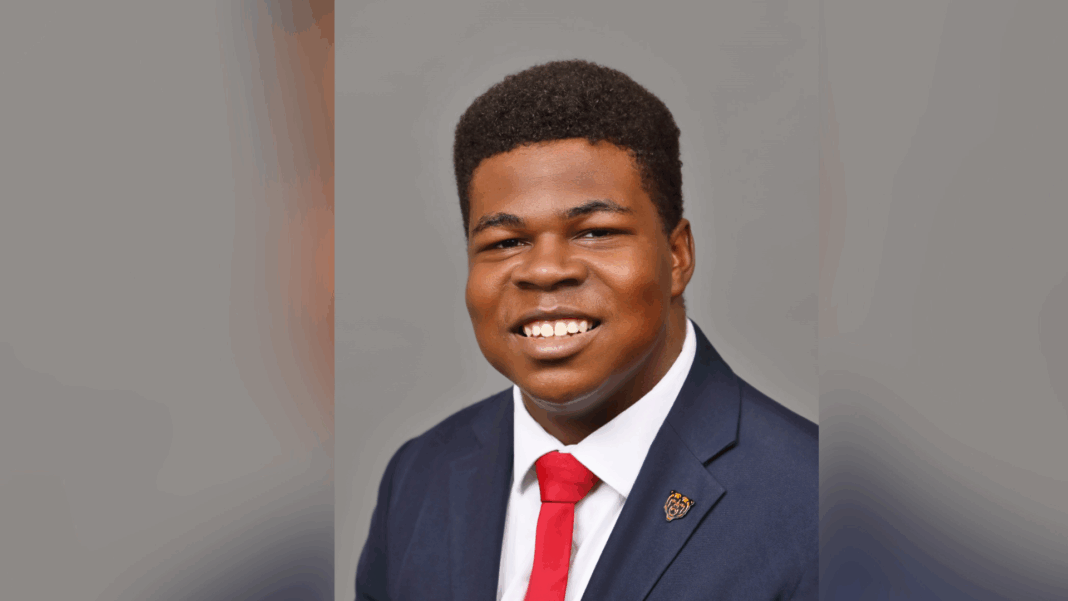 A young man in a suit and red tie smiles at the camera against a plain gray background.