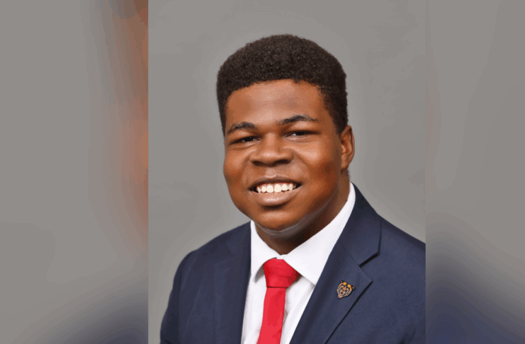 A young man in a suit and red tie smiles at the camera against a plain gray background.