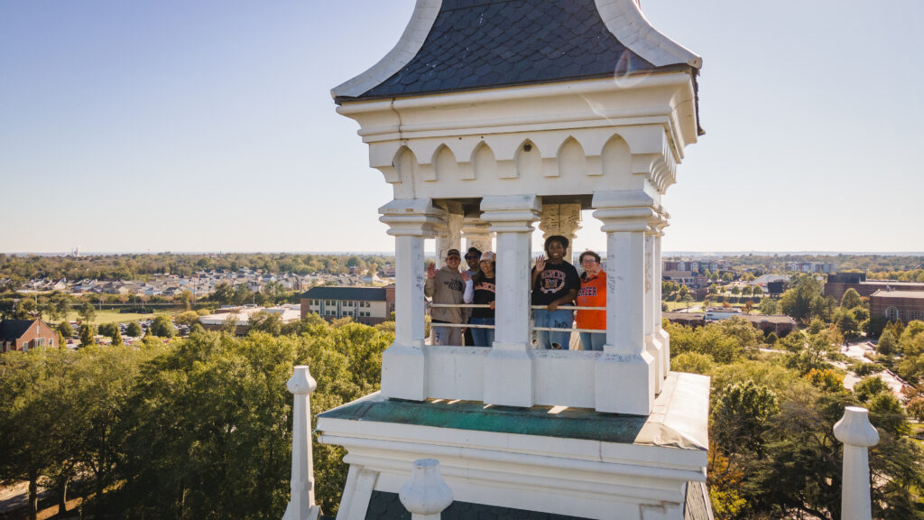 A group of people stand inside the upper level of a white bell tower overlooking a landscape of trees and buildings.