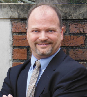 Man in a suit and tie smiles at the camera while standing in front of a brick wall outdoors.