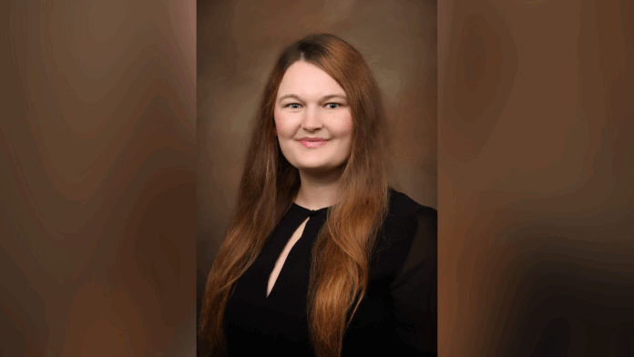 Woman with long brown hair wearing a black top, smiling at the camera against a brown gradient background.