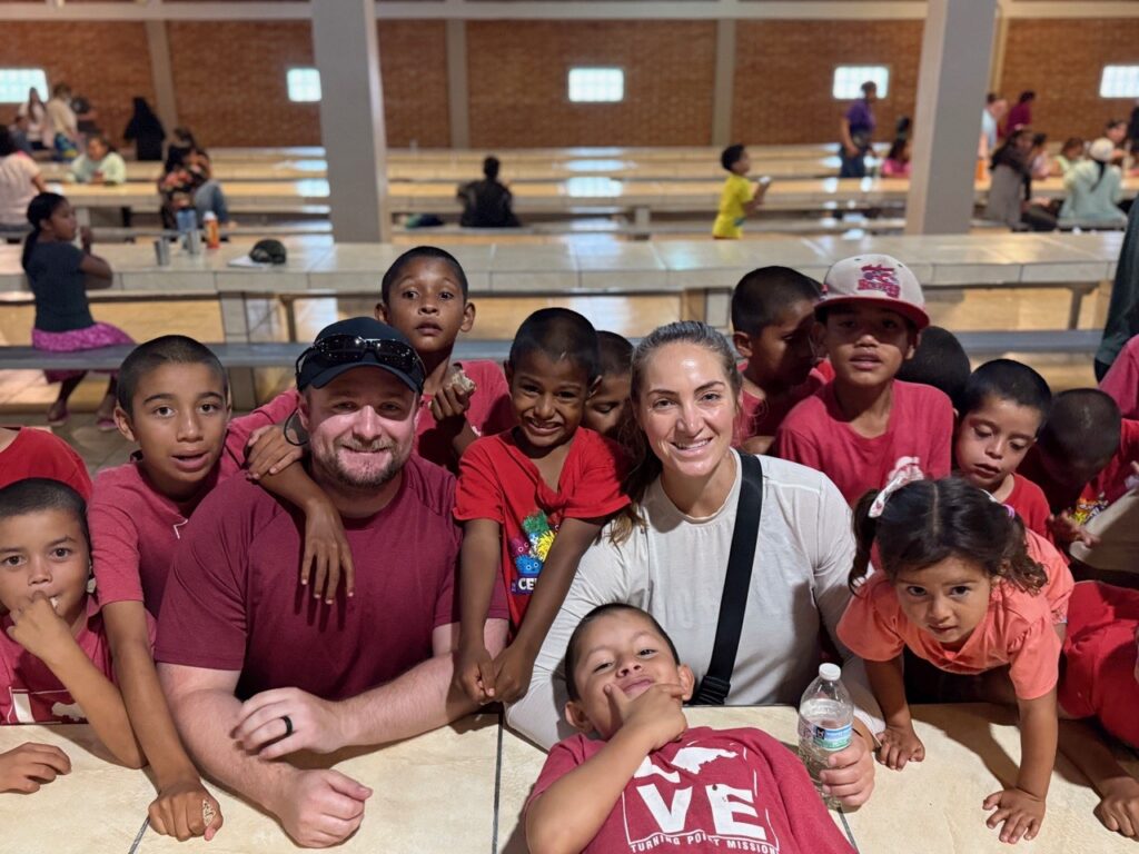Two adults sit at a table surrounded by smiling children in a large, well-lit hall.