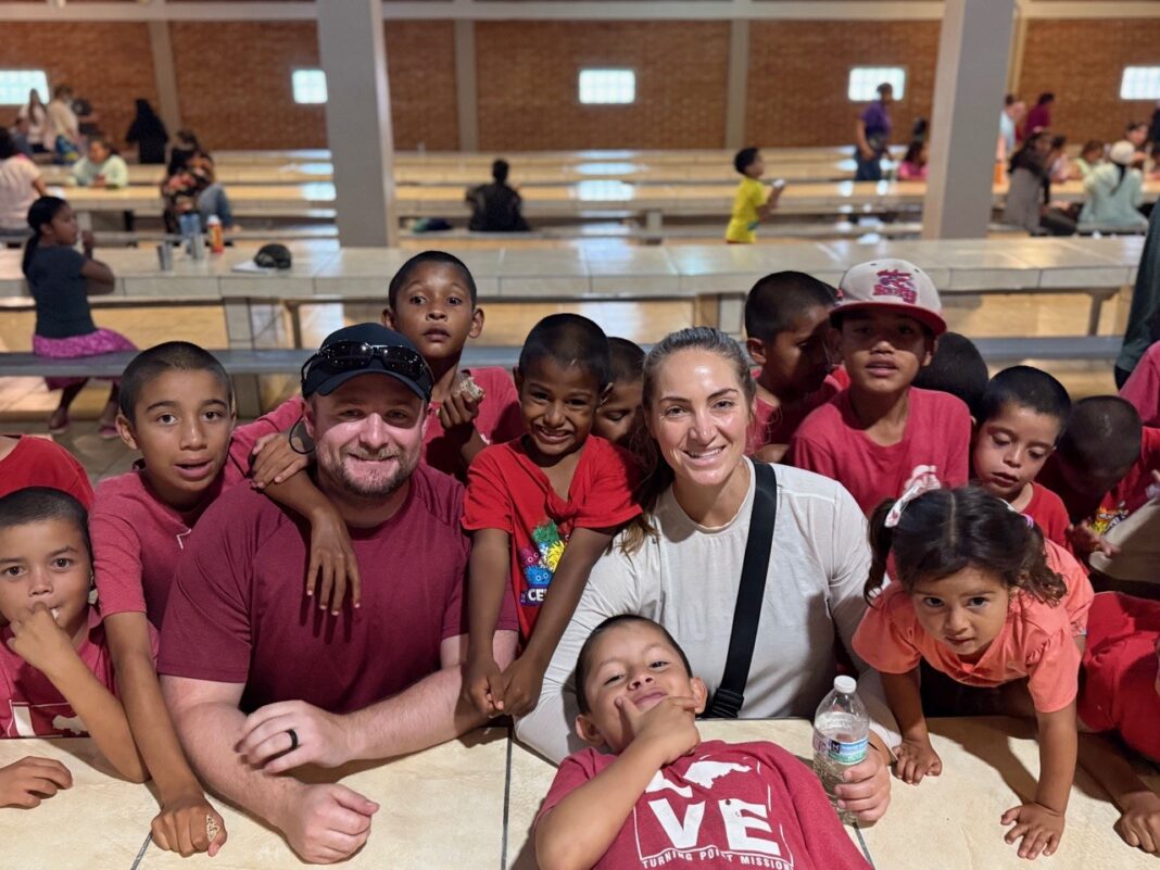 Two adults sit at a table surrounded by smiling children in a large, well-lit hall.