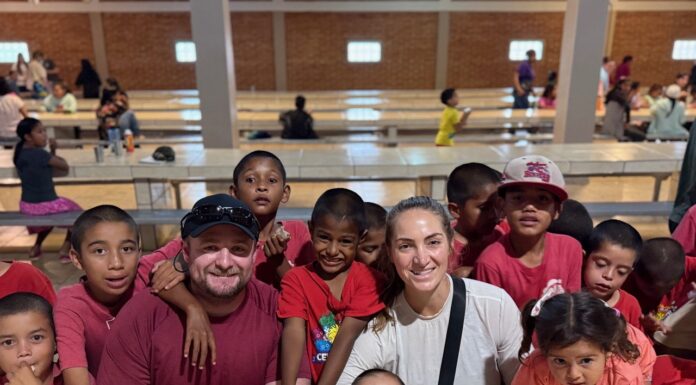 Mercer students set up medical clinic and telehealth connections in Honduras Two adults sit at a table surrounded by smiling children in a large, well-lit hall.