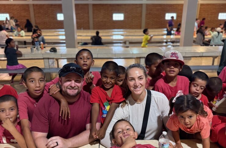 Two adults sit at a table surrounded by smiling children in a large, well-lit hall.