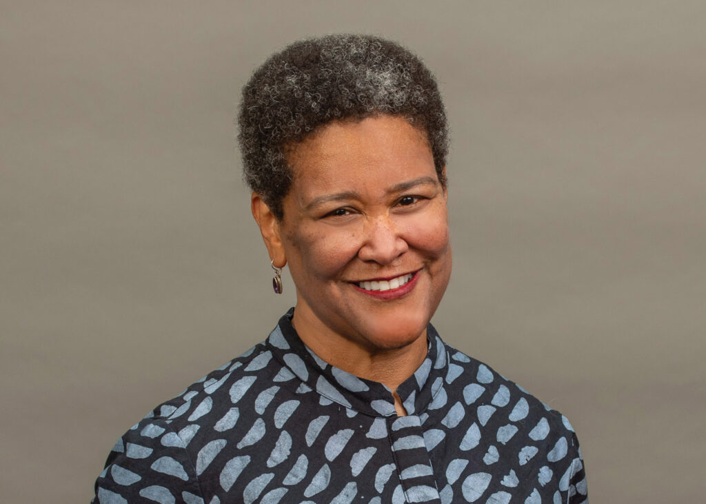 A woman with short curly hair and patterned shirt smiles in front of a plain, neutral background.