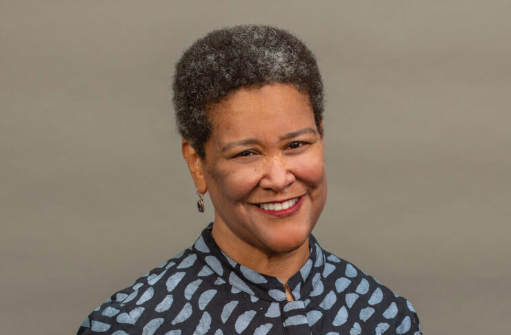 A woman with short curly hair and patterned shirt smiles in front of a plain, neutral background.