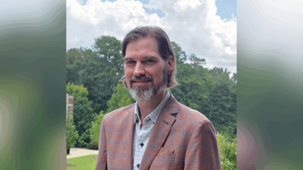 A man with brown hair and a beard wears a plaid jacket and stands outdoors in front of trees and a cloudy sky.