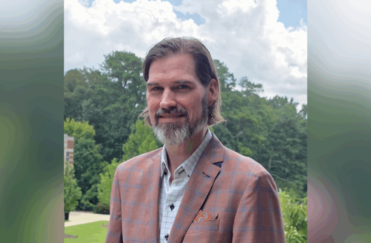 A man with brown hair and a beard wears a plaid jacket and stands outdoors in front of trees and a cloudy sky.