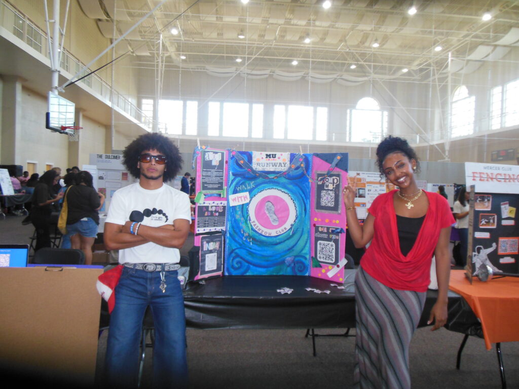 Two people stand beside a colorful display board at an indoor event with other booths in the background.
