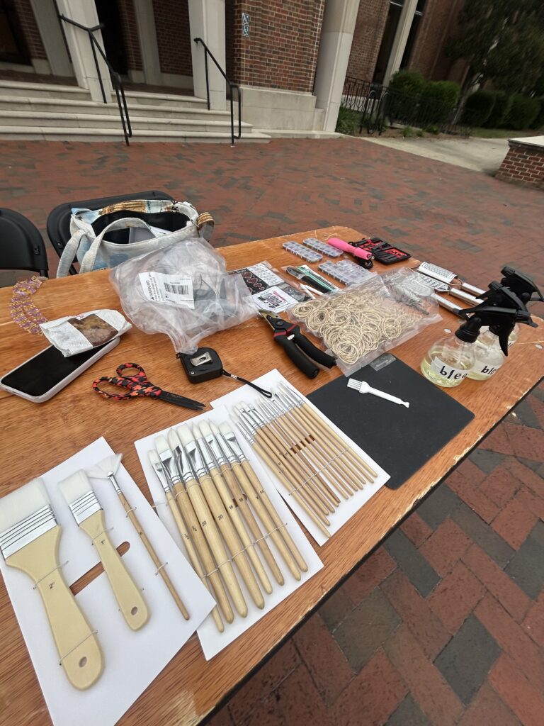 A table outdoors displays paintbrushes, rubber bands, tape, spray bottles, markers, and other assorted craft supplies.