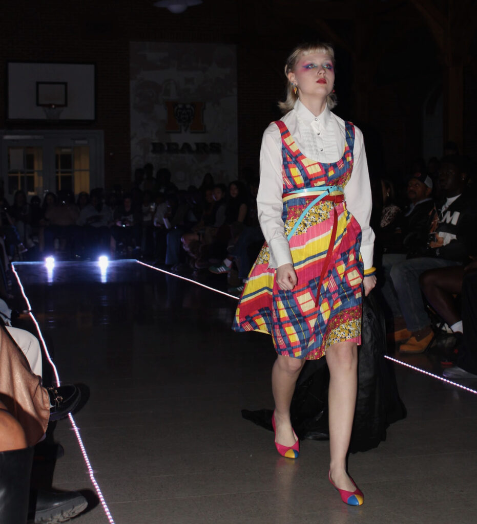 A model walks a runway wearing a colorful plaid dress over a white blouse with matching shoes.