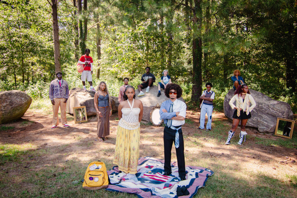 A group of people pose outdoors among trees and rocks, with mirrors and a blanket on the ground in the foreground.