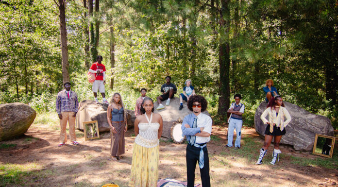 MU Runway wants students to ‘walk with confidence’ A group of people pose outdoors among trees and rocks, with mirrors and a blanket on the ground in the foreground.