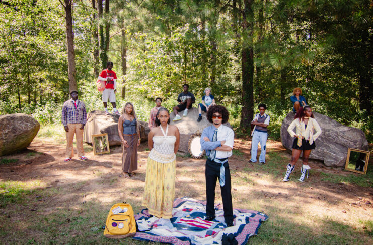 A group of people pose outdoors among trees and rocks, with mirrors and a blanket on the ground in the foreground.