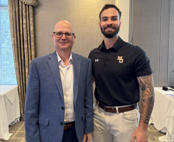 Two men stand side by side indoors; one in a blue blazer, the other in a black polo and light pants.