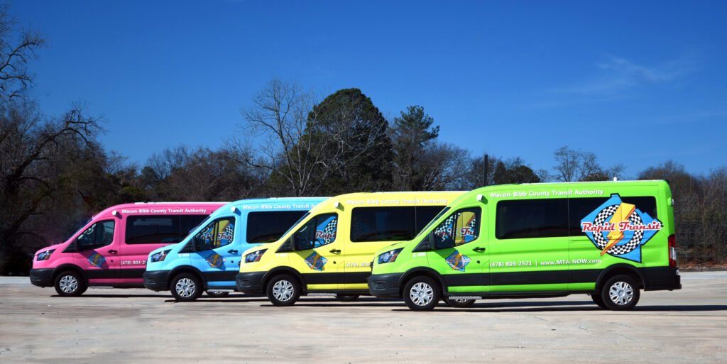 Four brightly colored Macon-Bibb County transit vans are parked side by side on a concrete lot under a clear sky.