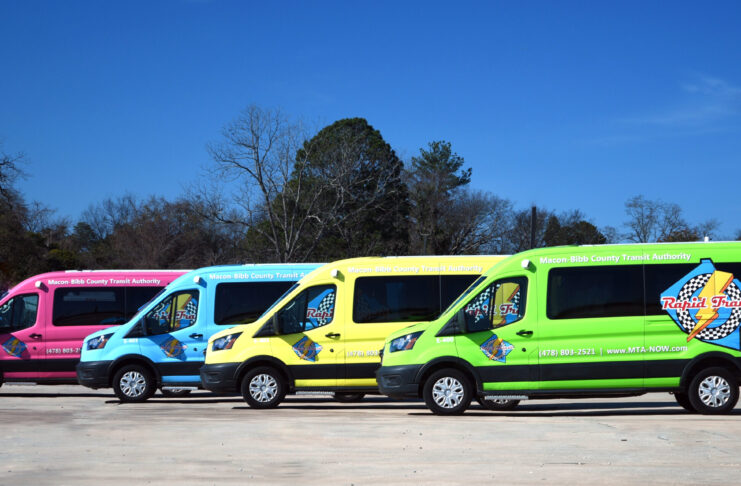Four brightly colored Macon-Bibb County transit vans are parked side by side on a concrete lot under a clear sky.