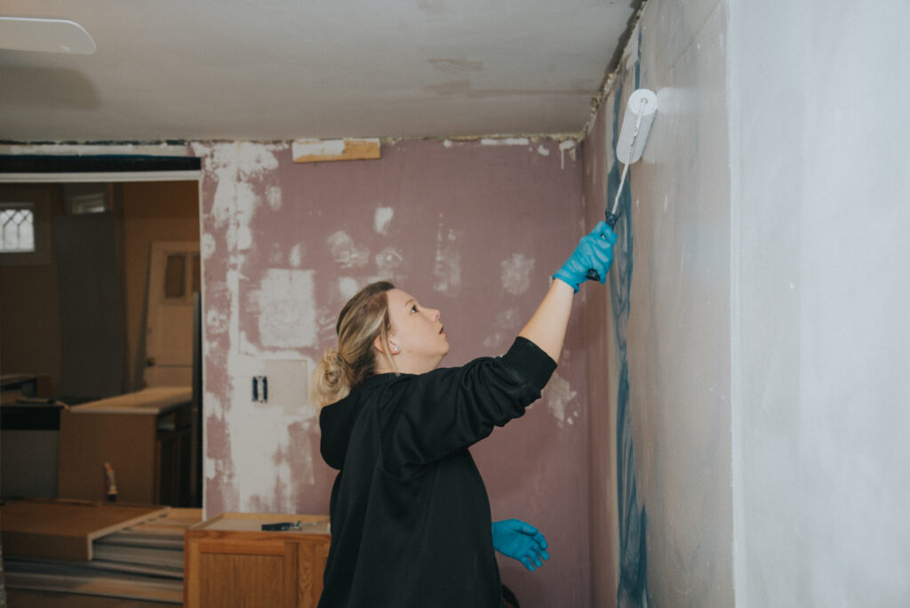 A Mercer student wearing gloves uses a paint roller to paint a wall in a partially renovated room.