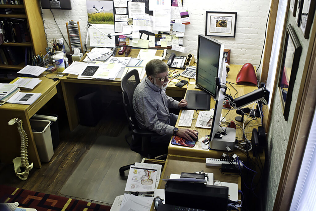 Man working on a computer in a cluttered office with papers, books, and anatomical diagrams on the desks around him.