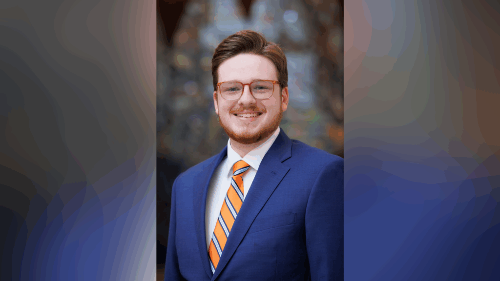 A man with glasses and a beard smiles, wearing a blue suit, white shirt, and orange-striped tie, against a blurred background.